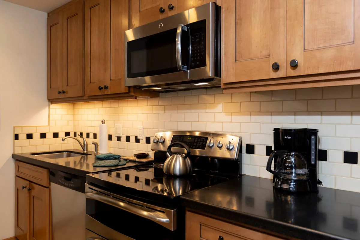 Here, the kitchen features stainless steel appliances, a black countertop, wood cabinetry, and essential small appliances like a kettle and coffee maker. The scene implies warmth and functionality, perfect for morning coffee routines or prepping a group meal.