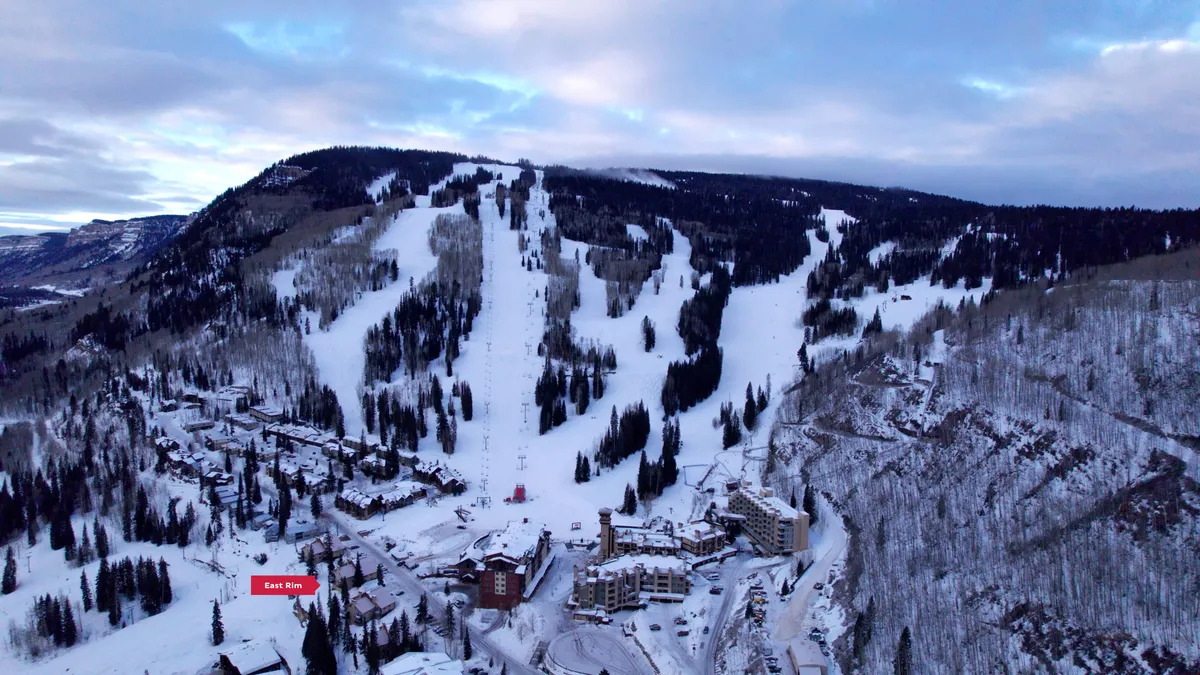 An exterior view of ski slopes weaving through pine trees with resort buildings nestled at the base. The snow-covered scene is peaceful yet active, evoking the thrill of sunrise skiing and the welcoming comfort of the resort after a day outdoors.