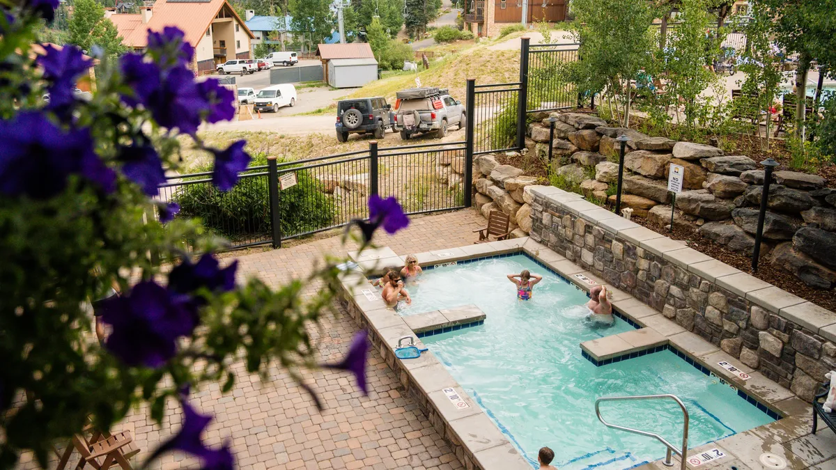 An inviting outdoor swimming pool is surrounded by blooming flowers and a rustic rock wall. People are enjoying the pool, some swimming and others relaxing poolside, while the setting sun casts a warm glow over the scene.