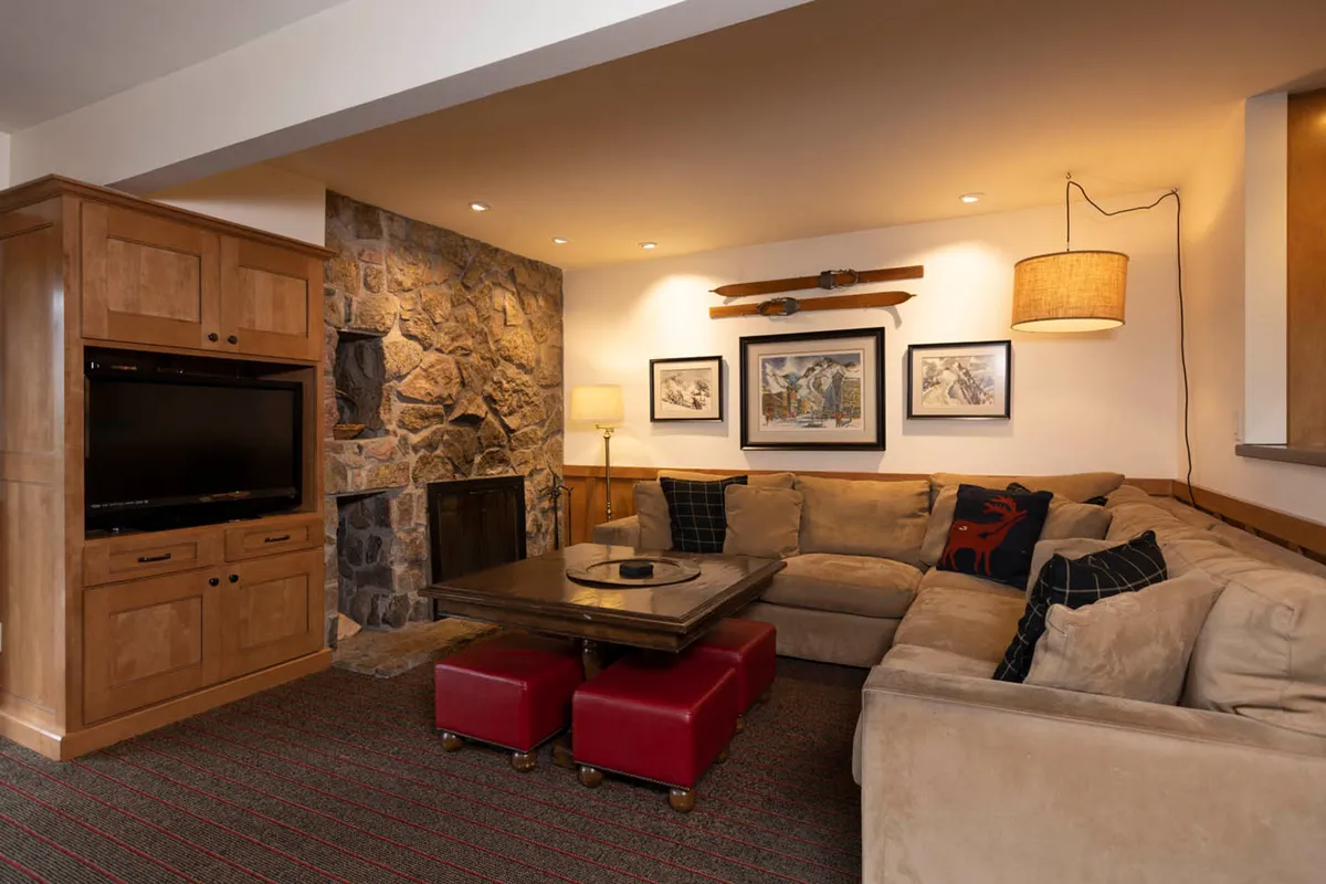 A spacious living room featuring a large sectional sofa arranged around a prominent stone fireplace. Red ottomans and a wooden entertainment unit add warmth, while framed artwork and wide windows create a welcoming atmosphere ideal for gatherings.
