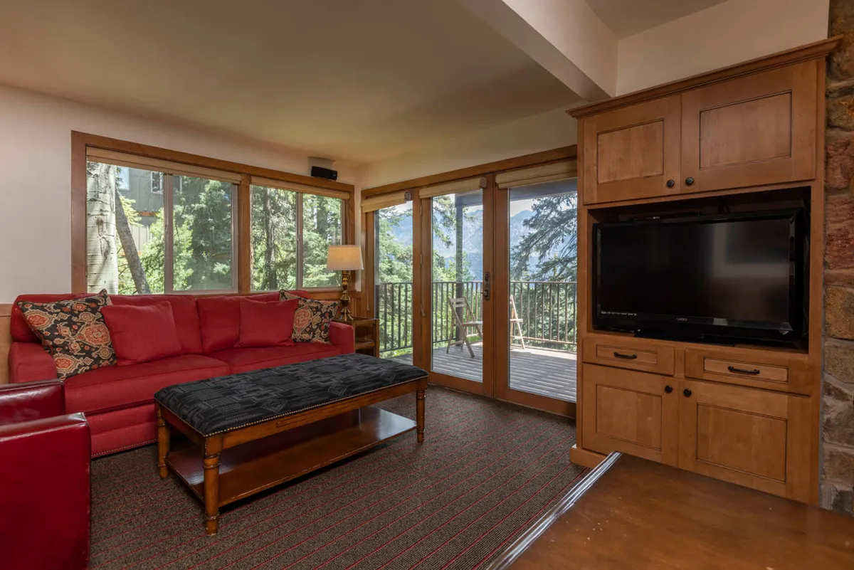 A cozy interior living room with a red sofa, wooden coffee table, large windows framing snowy outdoor views, and a television unit. The space invites relaxation after a day on the slopes, with hints of sunrise or snow scenes outside enhancing the connection between indoor comfort and outdoor adventure.