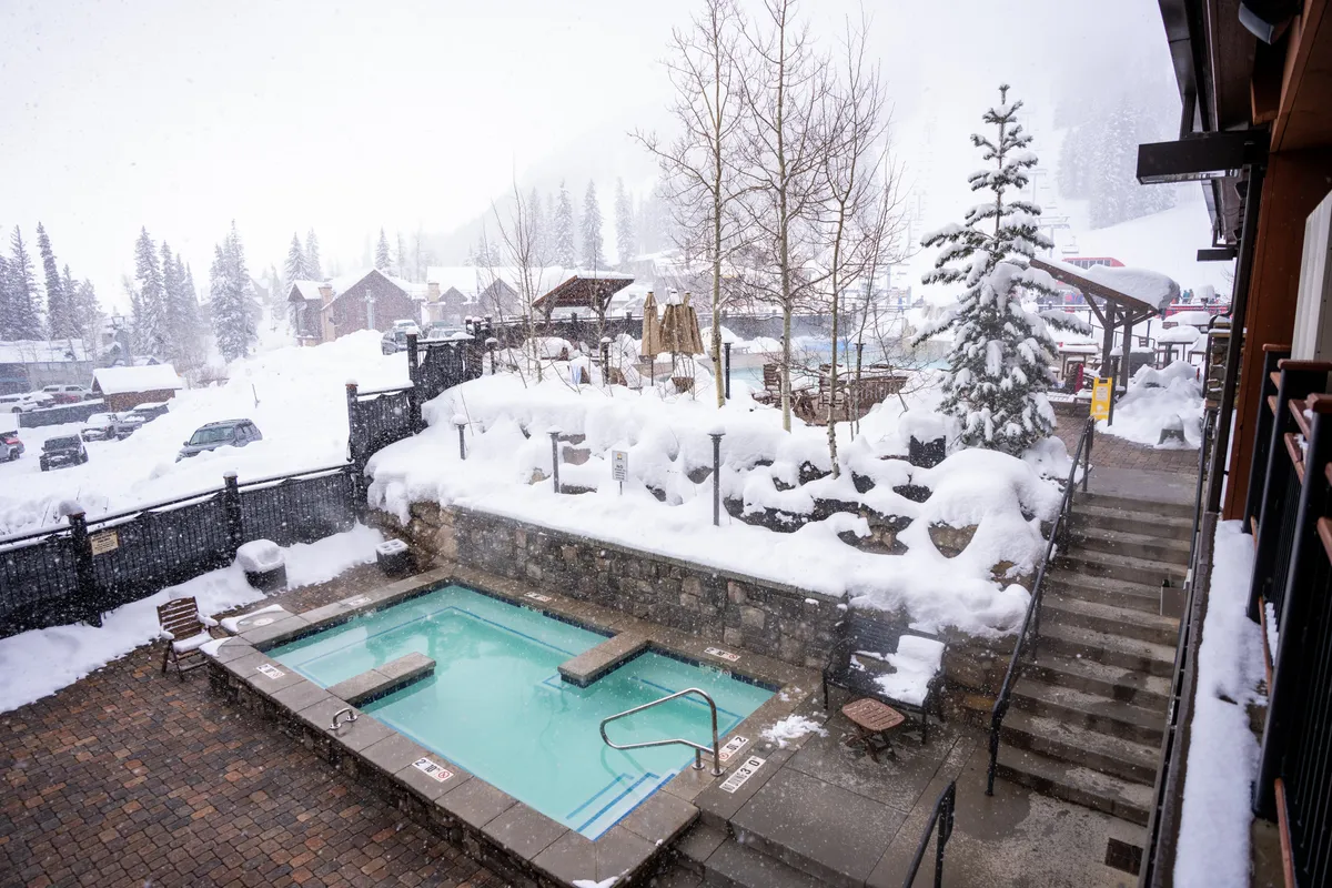 The image shows a steaming hot tub surrounded by snow, with tall trees and steps leading down to the cozy spot. The snowy landscape creates a tranquil setting, inviting guests to relax, socialize, and unwind together after a day of mountain activities.