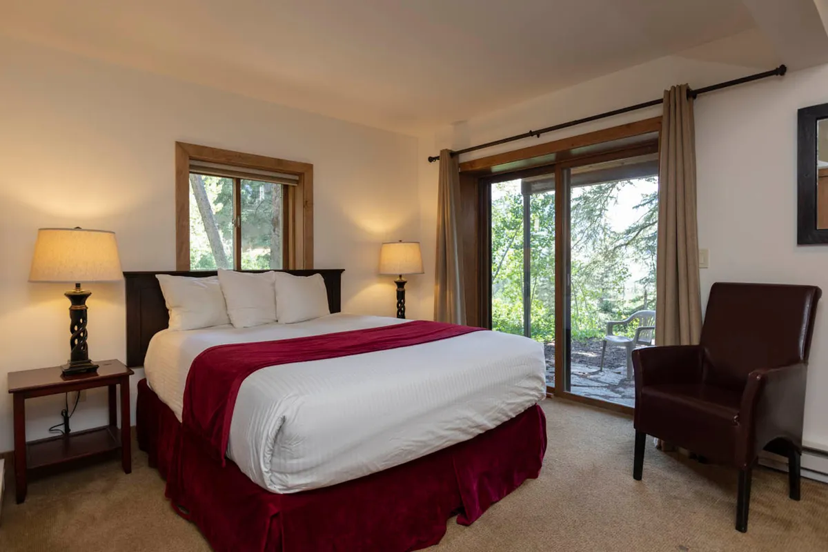 A tranquil bedroom with a plush bed featuring a red accent, large windows that let in natural light, an armchair positioned for relaxation, and soft lamp lighting creating a serene atmosphere.