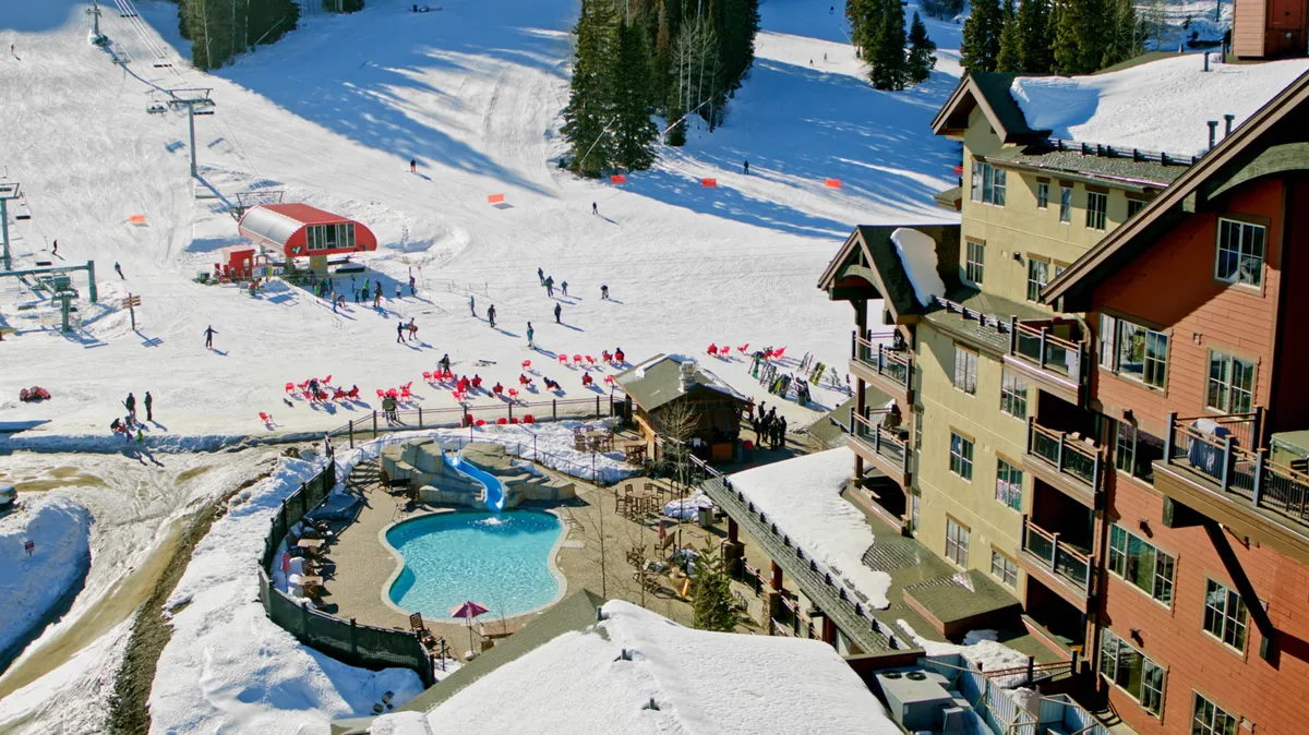 An exciting exterior view encompassing ski slopes, a swimming pool, ski lift, and resort buildings. The lively atmosphere suggests both winter and summer activities, with families skiing or snowboarding as well as relaxing by the pool. The scene captures the energy and diversity of resort amenities available to families just steps from their accommodation.