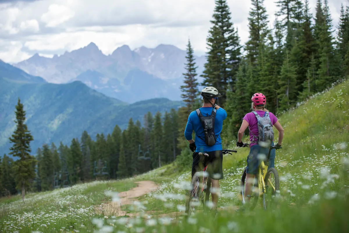 A dynamic outdoor scene featuring bikers on a trail lined with vibrant wildflowers, set against a backdrop of rolling mountains. The image evokes a sense of adventure and the joys of exploring nature, hinting at the hiking and warm-weather activities mentioned in the section. The community vibe among outdoor enthusiasts is palpable, making it ideal for adventurous families.