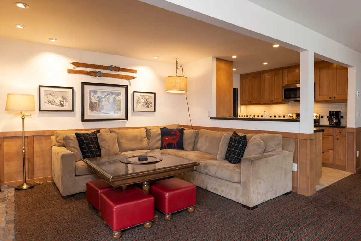 The image presents a spacious dining/living area with a large sectional sofa, wooden coffee table, bold red ottomans, and wall artwork. The setup exudes comfort and sociability, indicating a seamless transition from the kitchen to a communal dining or lounging space where groups can gather post-meal.